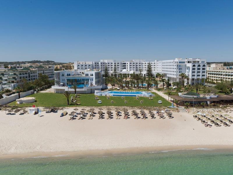 Hotel complex with pool, sun loungers, and direct access to sandy beach under clear sky.