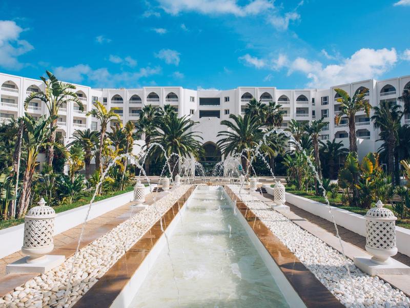 Luxury resort with white hotel, palm trees, and water feature under blue sky.