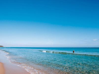 Strand mit hellem Sand, klarem blauem Wasser und wolkenlosem Himmel, einige Menschen im Wasser.