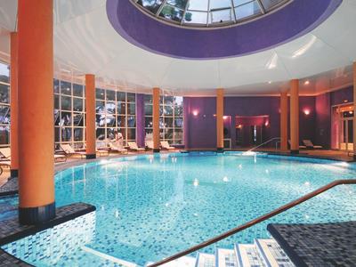 Großes Hallenbad mit runden Säulen, Glasdach und Liegestühlen am Beckenrand.