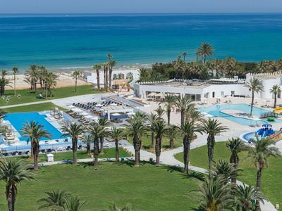 Strandhotel mit Pools, Palmen und grünen Rasenflächen am blauen Meer unter klarem Himmel.