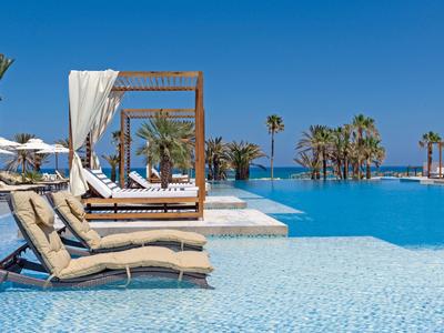 Área de piscina lujosa con tumbonas, camas con dosel y palmeras bajo un cielo azul claro.