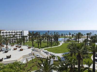 Hotelanlage mit Palmen, gepflegtem Rasen und Blick auf das Meer bei klarem Himmel.