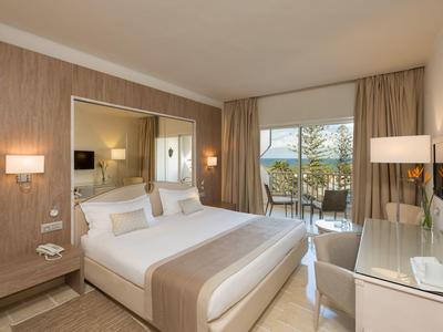 Helles Hotelzimmer mit Doppelbett, Holzvertäfelung und Balkon mit Meerblick.