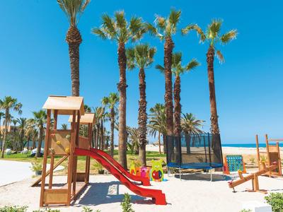 Spielplatz mit Rutsche und Trampolin auf weißem Sand, Palmen und blauer Himmel im Hintergrund.