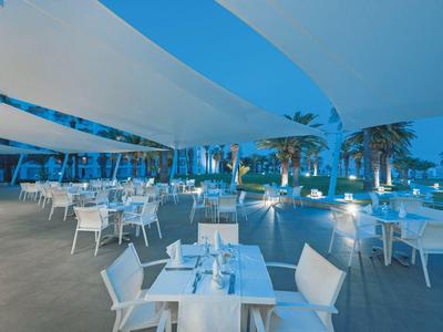 Moderner Außenbereich eines Restaurants mit weißen Tischen, Stühlen und bläulicher Beleuchtung, Palmen im Hintergrund.