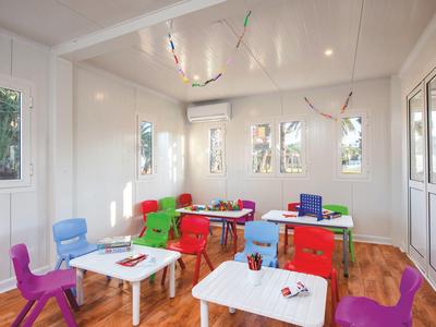 Buntes Klassenzimmer mit kleinen Tischen, bunten Stühlen und Holzfußboden, helle Wände und Fenster.