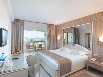 Helles Hotelzimmer mit Doppelbett, Balkon, Meerblick, Fernseher und moderner Einrichtung in Holz und Weiß.