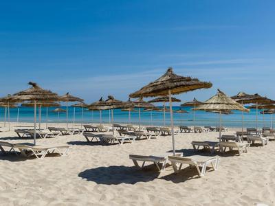 Strand mit hellem Sand, blauen Liegestühlen und Stroh-Sonnenschirmen unter klarem Himmel.
