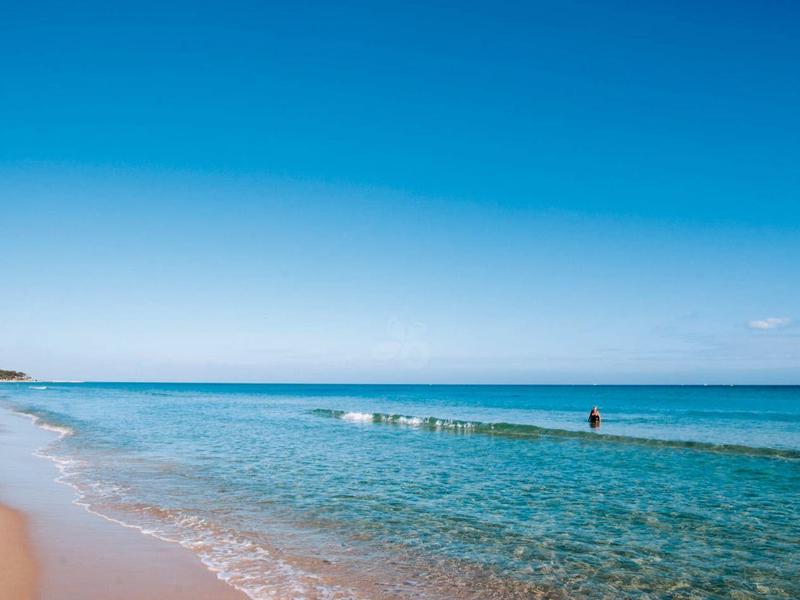 Strand mit hellem Sand, klarem blauem Wasser und wolkenlosem Himmel, einige Menschen im Wasser.