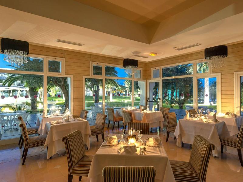 Elegantes Restaurant mit gedeckten Tischen, Kerzenlicht und großen Fenstern mit Blick auf Palmen.