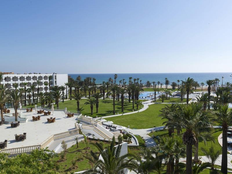 Hotelanlage mit Palmen, gepflegtem Rasen und Blick auf das Meer bei klarem Himmel.
