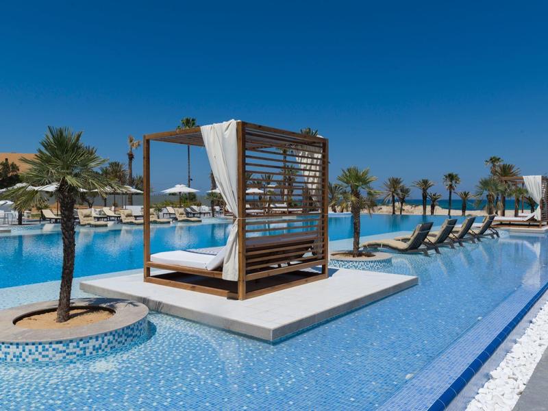 Moderner Pool mit Liegen, Palmen und Baldachin unter klarem, blauem Himmel an sonnigem Tag.