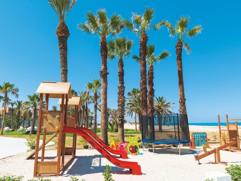Spielplatz mit Rutsche und Trampolin auf weißem Sand, Palmen und blauer Himmel im Hintergrund.