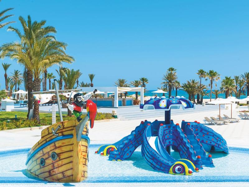 Kinder spielen auf einem Spielplatz mit einem Boot und einem Drachen, umgeben von Palmen und blauen Himmel.