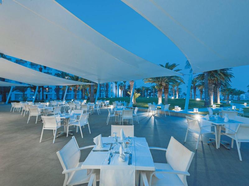 Moderner Außenbereich eines Restaurants mit weißen Tischen, Stühlen und bläulicher Beleuchtung, Palmen im Hintergrund.