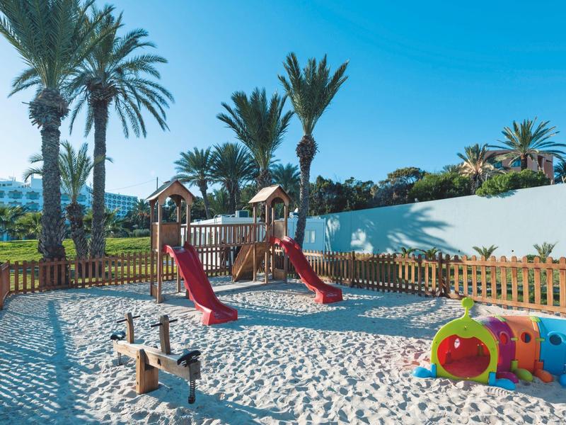 Kinderspielplatz mit Sand, Rutschen, Schaukelpferd und bunter Raupe, umgeben von Palmen und blauem Himmel.