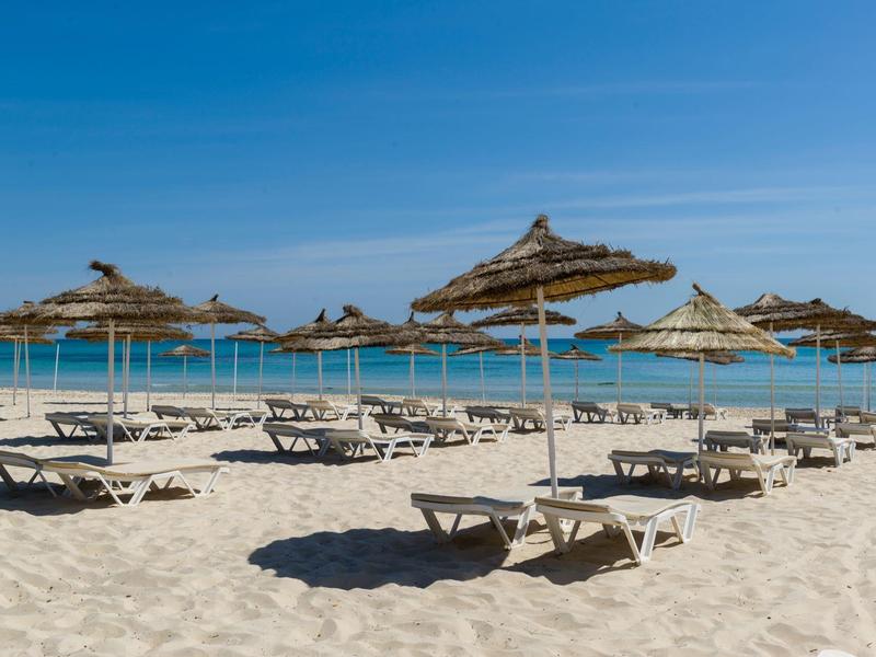 Strand mit hellem Sand, blauen Liegestühlen und Stroh-Sonnenschirmen unter klarem Himmel.