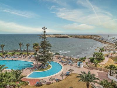 Blick auf einen Poolbereich und das Meer an einer Küstenanlage mit Palmen und Liegestühlen.