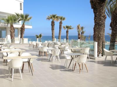 Leere Terrasse mit weißen Stühlen und Tischen, Palmen und Meerblick im Hintergrund.