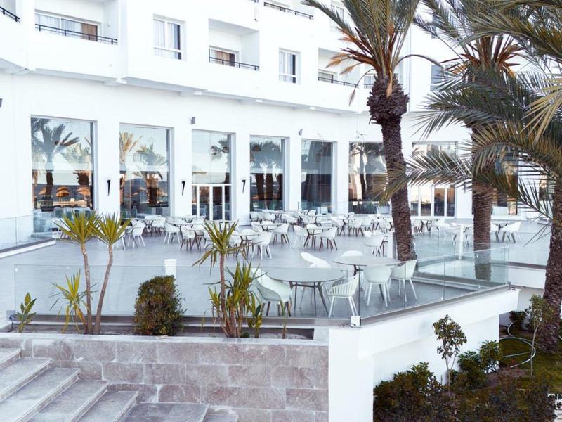 Ampia terrazza dell'hotel con tavoli, sedie e palme in una giornata soleggiata.