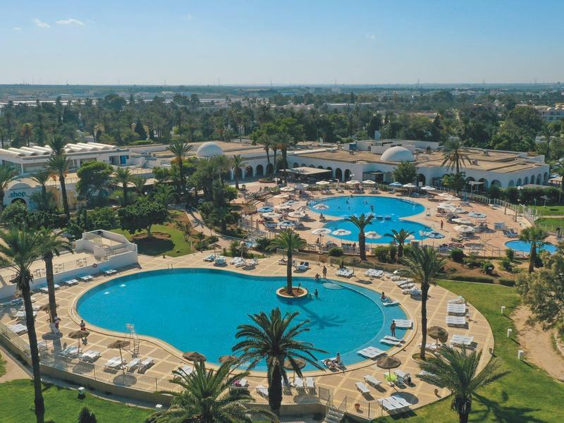 Grand espace piscine d'hôtel avec parasols, chaises longues et palmiers sous un ciel dégagé