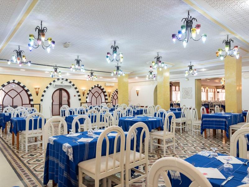Grande salle à manger avec nappes bleues et chaises blanches, lumineuse et accueillante.