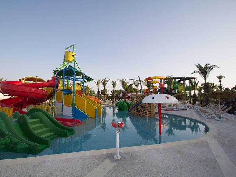 Parc aquatique coloré avec toboggans et champignon d'eau, entouré de palmiers et parasols.