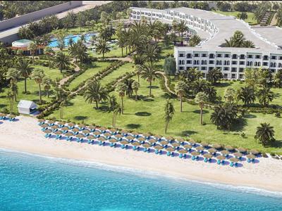 Complejo hotelero con edificios blancos, piscina, palmeras y playa con sombrillas junto al mar claro.