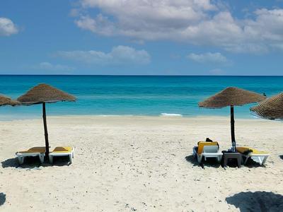 Strand mit weißem Sand, blauem Meer und drei Sonnenschirmen mit Liegestühlen darunter.