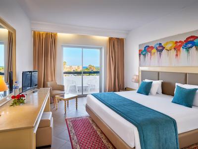 Bright hotel room with double bed, blue runner, balcony, and city view.