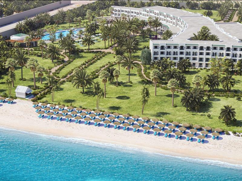 Complejo hotelero con edificios blancos, piscina, palmeras y playa con sombrillas junto al mar claro.