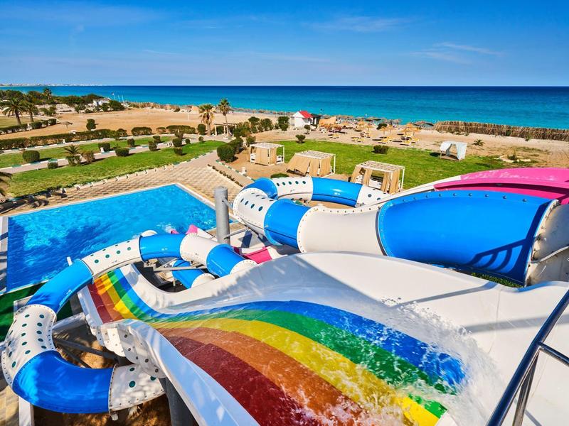 Piscina con tobogán de colores con vista a la playa y al mar bajo un cielo despejado.