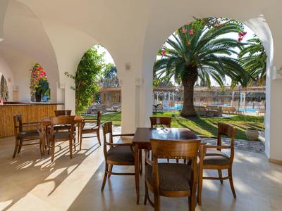 Pohodlná venkovní terasa s stoly a židlemi pod bílými oblouky, výhled na zahradu s palmou.