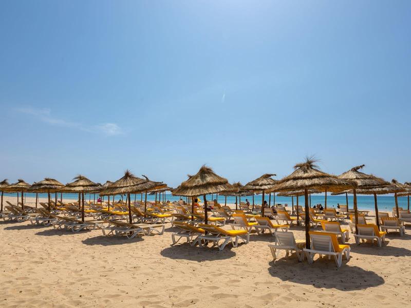 Pláž s řadami slámových slunečníků nad lehátky pod modrou oblohou