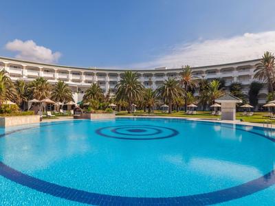 Groot hotel met tropische tuin en ronde pool onder blauwe lucht.