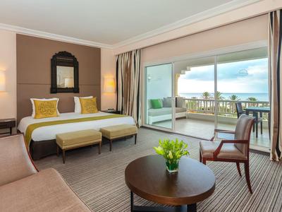 Elegant hotelkamer met groot bed, zithoek en zeezicht door grote glasramen.