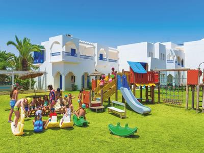 Kinder spielen auf einem bunten Spielplatz vor weißen Gebäuden mit blauen Akzenten unter blauem Himmel.