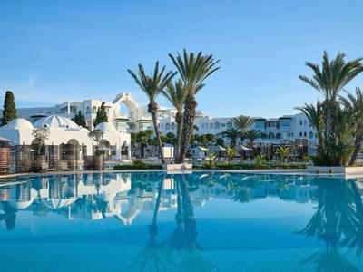 Piscina grande y limpia frente a un hotel blanco con palmeras bajo un cielo azul claro.
