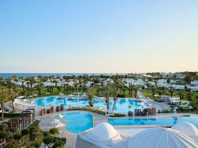 Vista de una gran piscina de hotel con piscinas azules, rodeada de palmeras y edificios blancos.