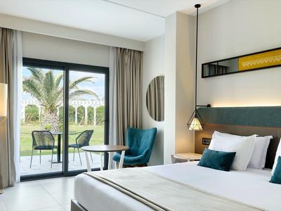 Habitación de hotel luminosa con cama doble, sillón y balcón con vista a palmeras.