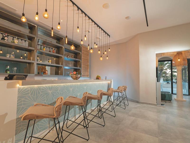 Área de bar de hotel moderna con mostrador iluminado, taburetes de madera y luces colgantes en interior luminoso.