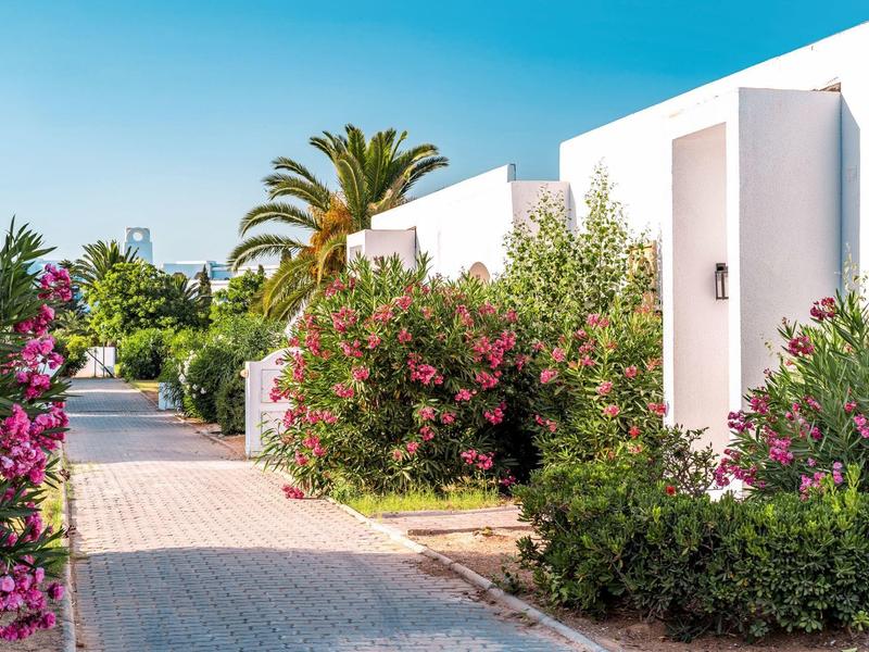 Edificio de hotel blanco con plantas verdes frondosas y flores rosas bajo un cielo azul.