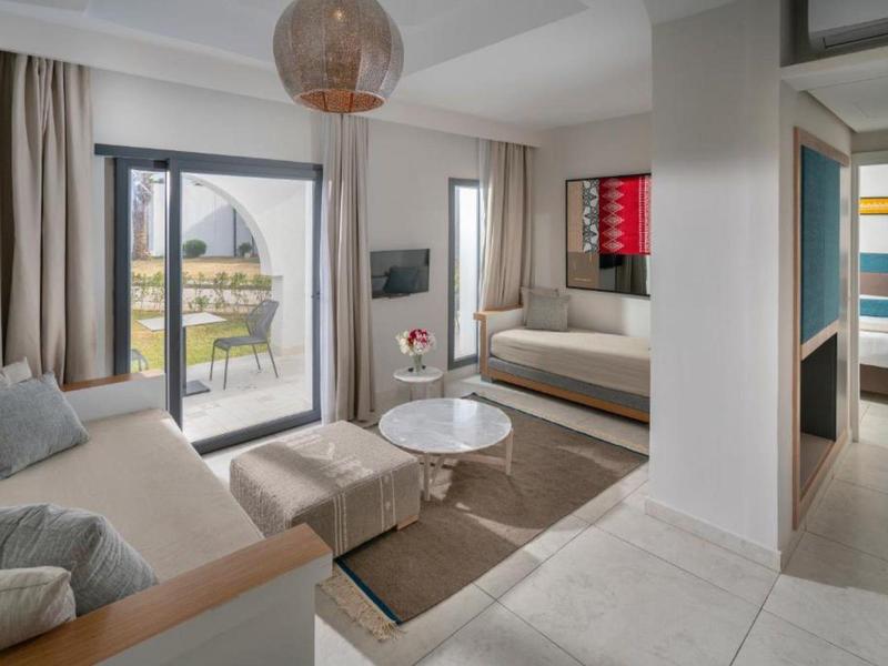 Sala de estar luminosa y moderna con sofá, sillón, alfombra y acceso a balcón en habitación de hotel.