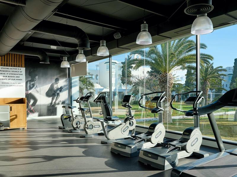Gimnasio moderno con equipos de entrenamiento frente a grandes ventanas y vista a palmeras.