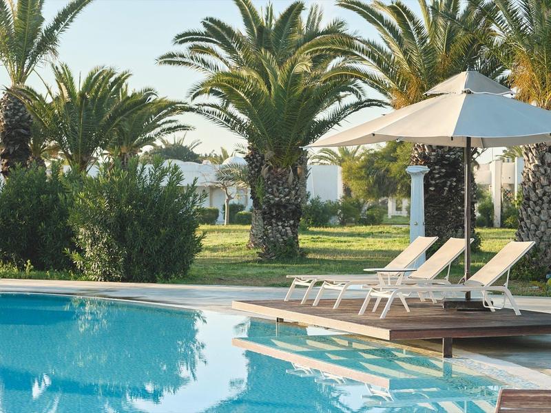 Área de piscina con tumbonas y sombrilla, rodeada de palmeras y paisaje verde.