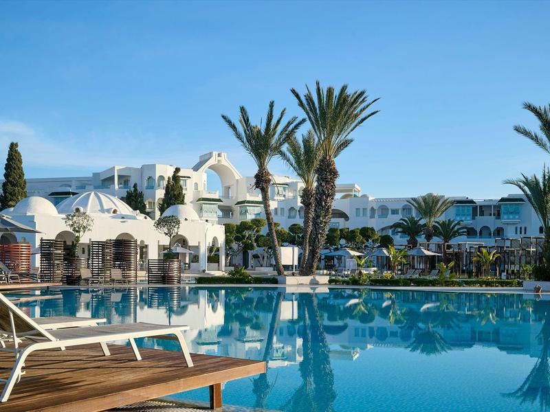 Un hotel con piscina, edificios blancos, palmeras y una tumbona de madera junto al agua.