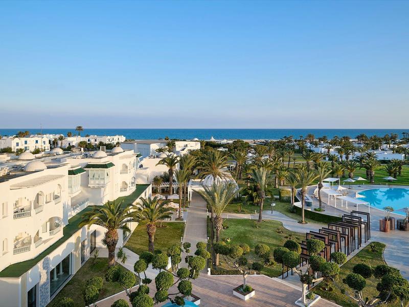 Vista de un complejo hotelero con piscina, palmeras y vista al mar bajo cielo despejado.