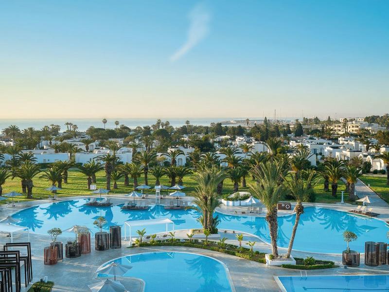 Gran piscina con palmeras y edificios blancos en un destino vacacional soleado.