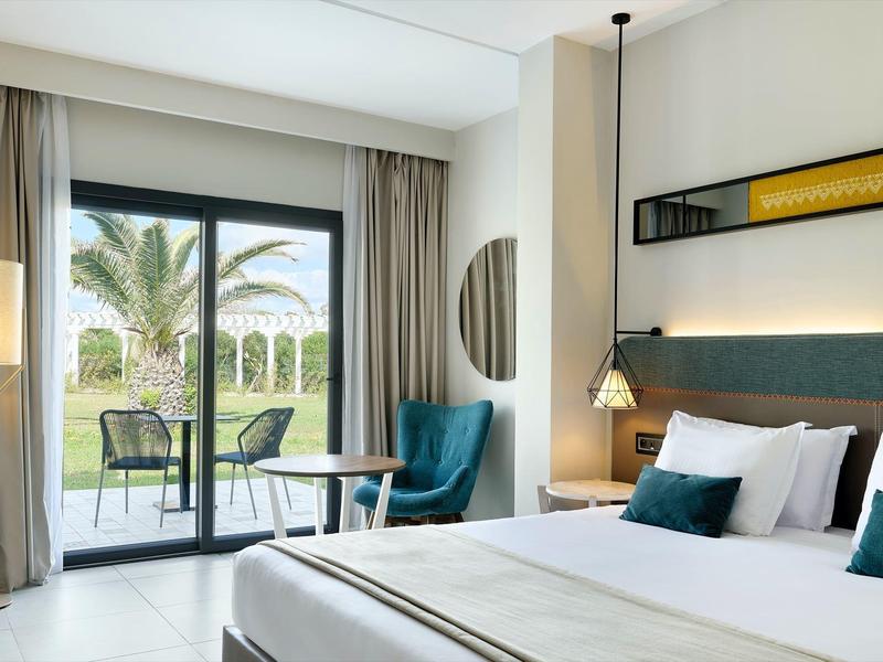 Habitación de hotel luminosa con cama doble, sillón y balcón con vista a palmeras.